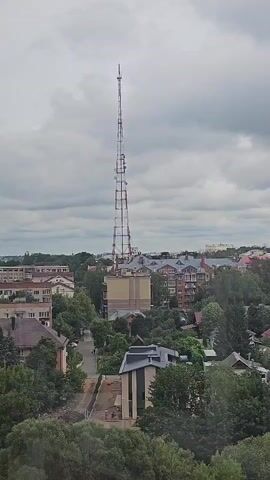 A high-angle, medium shot shows a tall, lattice telecommunications tower with a conical antenna on top. The tower is located in a town, surrounded by trees and buildings. The sky is overcast with gray clouds. The tower is white and red, with horizontal white bands and red vertical supports. There are several antenna dishes mounted on the tower at different levels. The buildings are of various sizes and architectural styles, with red tile roofs. Some of the buildings appear to be residential. In the foreground, lush green trees obscure the bottom of the scene. The overall impression is that of a typical urban or suburban landscape under a cloudy sky.