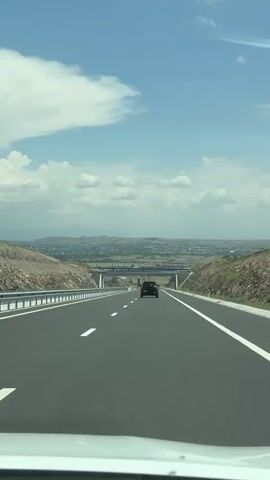 This media file depicts a forward-facing view from inside a vehicle traveling on a highway. The highway is modern, with clear white lane markings and a metal guardrail along the left side. The road is flanked by rocky, arid terrain with sparse vegetation on either side. In the distance, a city or town is visible spread across a valley, with mountains forming the backdrop. A blue bridge spans the highway ahead, and a dark-colored SUV is driving in the same direction, further down the road. The sky is a bright blue with scattered white clouds, suggesting a clear and sunny day. The overall impression is one of travel and movement through a scenic, possibly mountainous, landscape. The location is identified as Aghdzk, Armenia. There is no visible text in the media file.