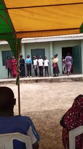 From a seated perspective under a green-and-yellow canopy, the video shows a group of nine people of varying ages and skin tones standing on a concrete platform in front of a pale green building with dark green barred windows and a doorway. The people are dressed in formal and casual attire. The building sits on a gravel ground, and people are milling about near the entrance. One woman in a yellow jacket and dark hat speaks into a microphone while holding a brown bag.  The scene appears to be an outdoor event or ceremony.