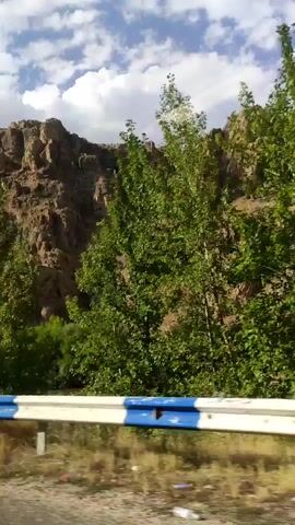 A video captures a roadside view of a rocky mountain covered with green trees. A guardrail with blue and white stripes runs along the bottom of the frame, separating the road from the mountainside. The sky above is partly cloudy with patches of blue. The camera appears to be moving, giving a sense of travel.