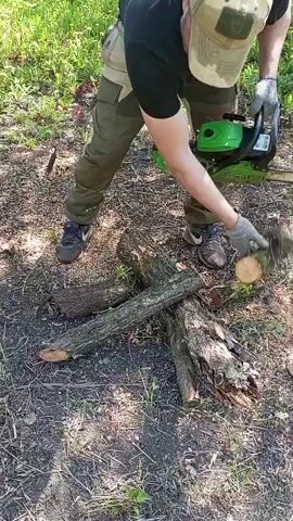 A person wearing a black shirt, beige pants, and a light-colored cap is using a green and black chainsaw to cut a log in a grassy outdoor area. The person is also wearing gloves and uses both hands to operate the chainsaw. The log is resting on other pieces of wood on the ground. The ground is covered in dry leaves and grass.