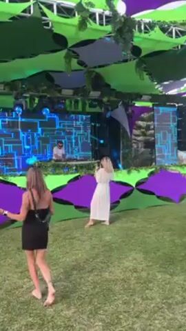 The video shows a live DJ set at an outdoor event. The DJ, wearing a white shirt and sunglasses, is performing on a stage with a large screen displaying a blue and white grid-like pattern. Two women stand in the foreground on a grassy field. One is wearing a short black dress and the other a long white dress. The stage is decorated with green and purple accents, and there are hanging green decorations above. The music is loud and electronic.