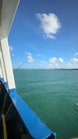 The video shows a view from a boat on a sunny day with a clear blue sky and scattered white clouds. The water is a turquoise green and appears to be slightly choppy. In the distance, a shoreline with some industrial structures and buildings is visible. A fishing rod is extended from the side of the boat, suggesting someone might be fishing. The sun is bright and reflects off the water.