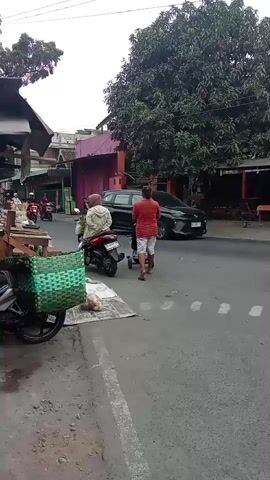 In a daylight outdoor scene, several people on motorcycles are visible on a street. A black SUV is parked on the right side of the road. On the left side of the road, a motorcycle with a green basket attached is parked, and next to it, there are some items on the ground. Two individuals, one wearing a light-colored jacket and the other in a red and white striped shirt, are standing near a motorcycle. Another person is walking away from the camera toward the street. The environment appears to be a marketplace or a bustling street with shops or buildings lining the road and trees providing shade.
