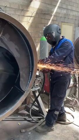 A worker is grinding a large, rusty metal ring in a workshop. He is wearing a blue uniform and a black welding helmet, which is lit up from the inside. The grinder sends off a bright shower of sparks. The ring is held in place by a metal support structure. The workshop has cinder block walls and is lit by natural light.