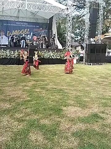 In a wide shot, on a lawn, four women are dancing in front of an elevated stage on a bright day. The dancers are all wearing reddish brown clothing, hair accessories, and holding red fans. They are dancing in coordinated movements. The stage is behind a line of black planters filled with white flowers. There are musicians visible on the stage along with a large banner that has "PENTAS SENI DALAM RANGKA HUT KAB. SRAGEN KE-280" and "DJADOEL Festival" written on it. A large speaker tower is next to the stage. A few people are standing around the perimeter. The lawn is green. The sky is a pale blue with some clouds.
