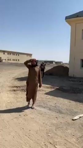 The video depicts a dusty outdoor setting, seemingly in a rural or arid environment. It features several men dressed in traditional Middle Eastern clothing.

At the beginning, a man in a brown robe is walking away from the camera, shielding his eyes from the sun with his hand. Two other men are walking behind him.

The scene then shifts to show a man with a rifle slung over his shoulder, standing near a beige building.

Next, two men are shown facing the camera. Both have beards and are wearing traditional clothing. The man on the left has a scarf around his neck and vest.

Finally, the video ends with a man walking away from the camera past rubble and debris. In the distance, there is a long concrete wall and mountains.