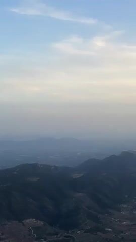 The video shows an aerial view of mountains and a valley. The sky is a gradient of light blue at the top, fading into a hazy white as it approaches the horizon. Wispy clouds are scattered across the sky. In the foreground, dark, rugged mountain peaks are visible, with some lighter colored patches suggesting vegetation or rocky outcrops. Beyond the foreground mountains, the landscape recedes into a hazy, blue-toned valley dotted with what appear to be buildings or settlements. The overall impression is of a vast, expansive landscape seen from a high vantage point, possibly from an aircraft. There are no people or distinct objects visible, only the natural environment. The lighting suggests it could be dawn or dusk, due to the soft light and the hazy atmosphere.