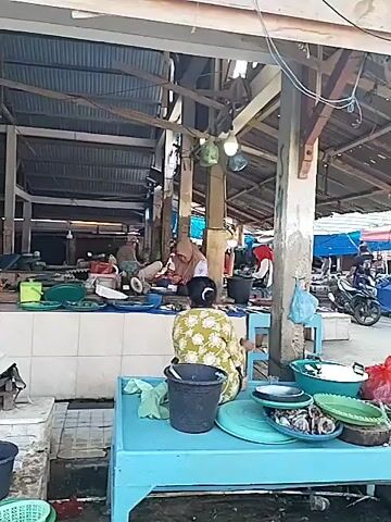 The video shows a fish market stall with two women working behind a counter. The woman on the right is wearing a green and white patterned shirt and appears to be seated and looking down at something. The woman on the left is wearing a purple headscarf and a long-sleeved shirt and is also seated. The counter is lined with white tiles and has various items on it, including scales, bowls, and what appear to be fish. In front of the counter, there are several white coolers or boxes stacked on top of each other, some with buckets on top. One cooler has the word "NIR" written on it in black marker. The market appears to be covered by a roof supported by wooden beams and metal poles. Some lights are visible hanging from the ceiling. The overall environment seems to be an open-air or semi-open market.