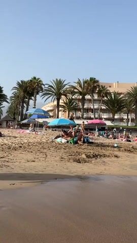 The video pans across a sandy beach with palm trees in the background, and a resort or hotel building behind the palm trees. There are many people on the beach, some are sunbathing under umbrellas and on towels, while others are in the shallow water. The water is calm and clear, with small waves lapping at the shore. The sky is clear and blue. The video starts with a close-up of the wet sand at the water's edge, then pulls back to reveal the entire beach scene. The camera continues to pan from left to right, showing more of the beach and the people on it. In the distance, a hill or mountain can be seen. There are children playing in the water, and adults are swimming and relaxing. Some people are walking along the beach, and others are sitting under umbrellas. The video ends with a view of the beach stretching out to the horizon.