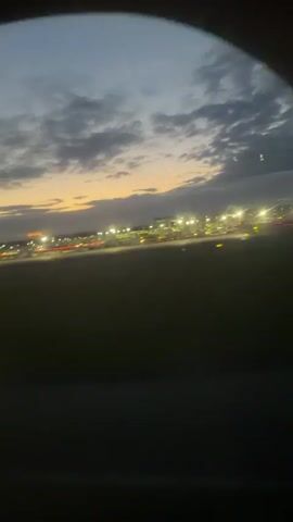 The video shows a view from an airplane window at dusk. The sky is dark with clouds, and the sun is setting on the horizon, casting an orange glow. Below, an airport runway is visible with many lights and a control tower. The airplane appears to be taxiing or just taking off.