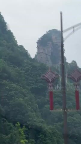 The video shows a roadside view of a mountain landscape. The foreground features decorative streetlights with red embellishments. The middle ground consists of a lush, green mountainside. A white car and a blue and white bus with the license plate ALY127 drive along the road. In the background, distinctive pillar-like rock formations rise into the misty sky. The overall impression is a scenic view of a natural landscape.