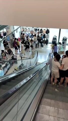 The video shows a busy department store from an elevated perspective, looking down at escalators and the shopping floor. Numerous people are visible, some on the escalators and others browsing merchandise on the floor below. The merchandise appears to be clothing and accessories, with racks of apparel and displays of bags. Several individuals are interacting with store employees, possibly at checkout counters or service desks. The environment suggests a well-lit retail space with multiple levels. The overall impression is one of active commerce and customer engagement.