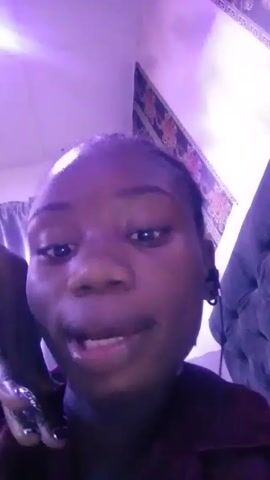 The video shows a young Black woman speaking to the camera. She is wearing a dark-colored shirt and has short hair slicked back. The lighting is poor, with a purple hue. Behind her, there is a wall hanging with floral patterns and a dark object, possibly a piece of furniture or clothing. She moves her neck slightly while she speaks, suggesting she may be uncomfortable or stretching. The room is dimly lit and looks like a personal space. The focus remains on her face throughout the short video.