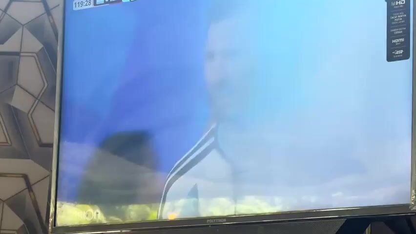 The video shows a wide shot of a soccer match being played on a green field in a stadium filled with spectators. The scoreboard at the top left of the screen indicates that the game is in its 119th minute, with Argentina (ARG) leading Switzerland (SUI) 2-1. Players in red and white uniforms are scattered across the field, engaged in play. A referee in a yellow shirt is also visible on the field. Advertising banners with the "aramco" logo are displayed along the sideline. The screen itself is a flat-panel television, mounted against a decorative wall.