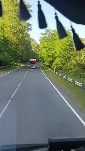 This video captures a view from inside a vehicle traveling on a paved road, likely in Ijevan, Armenia. The road curves to the right, flanked by lush green trees and foliage on both sides. A guardrail runs along the right side of the road, indicating a potential drop or embankment. Sunlight filters through the trees, casting dappled shadows on the asphalt, suggesting a clear day.

In the distance, a red truck with a white cargo area is visible, also traveling on the same road. The truck appears to be a utility vehicle or a transport truck. The camera is positioned to capture the road ahead and the surrounding natural environment. Several decorative tassels hang from the top of the vehicle's interior, obscuring small parts of the sky and trees. The overall mood is serene and suggests a leisurely drive through a scenic, wooded area. The time of day appears to be morning or early afternoon, given the bright sunlight. There is no visible text in the video.