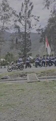 A group of people in uniform, possibly a marching band or a parade unit, is walking in formation on a paved road. They are wearing blue shirts and dark pants, and many are carrying instruments or equipment. A motorcycle is parked on the side of the road, and a flag with red and white stripes, likely the Indonesian flag, is visible. The background shows a natural landscape with trees and hills. The weather appears overcast.