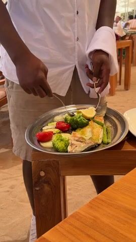A waiter wearing a white shirt and beige shorts stands at a wooden table, holding a metal platter with a cooked fish, broccoli, tomatoes, asparagus, and a slice of lemon. He is using a fork to arrange the lemon slice on the fish. Another person's hands are visible in the foreground, holding a fork and knife. The background shows an outdoor restaurant setting with wooden tables and chairs, umbrellas, and other diners. The ground appears to be sandy.
