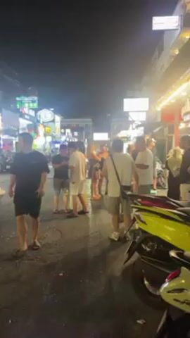 The video shows a busy street scene at night. Several people are standing and talking in small groups, illuminated by the bright lights of nearby shops and signs.  One person is sitting on a small stool. The street surface appears wet or damp. Several motorcycles are parked along the side of the road.  The scene suggests a lively atmosphere with people socializing in an urban setting.