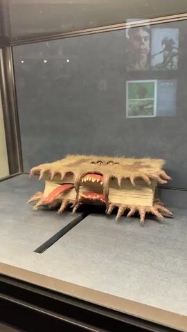 A short video shows a prop of the "Monster Book of Monsters" from the Harry Potter franchise, displayed in a glass case. The book has fur-like material covering it, with multiple eyes and a mouth that opens and closes. The book also has a tongue and what appear to be claw-like appendages on the edges. In the background, there are pictures on the wall, possibly of characters or creatures from the Harry Potter series.