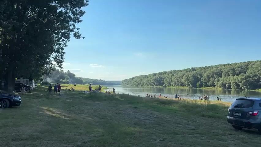 A wide shot shows a group of people gathered on the grassy bank of a wide river. The river is bordered by dense green forest on both sides, with a clear blue sky overhead. Several vehicles, including a black car and a dark blue SUV, are parked on the grass. Some people are standing, while others are sitting or lying down. A few people appear to be in or near the water. Smoke rises from what looks like a campfire in the background on the left side of the frame. The overall atmosphere appears to be relaxed and casual, suggesting a day of outdoor recreation.