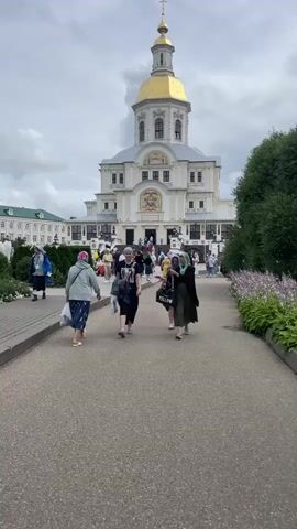 In front of a large, ornate white building with a golden dome, people walk on a paved path. The building appears to be a church or monastery. Some of the people are wearing headscarves. The sky is cloudy. There are trees and flowers on the right side of the path. Some people are carrying bags. The camera is positioned at a medium height, looking forward towards the building. The scene is well-lit, suggesting it is daytime.