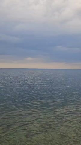 The video shows a view of the ocean on a cloudy day. The water is calm, with gentle ripples visible on the surface. The sky is overcast with a mix of gray and white clouds, and a narrow band of yellowish light is visible along the horizon. A single sailboat is visible on the left side of the frame in the distance. The water near the shore is clear enough to see the bottom, where light-colored rocks or sand are visible.
