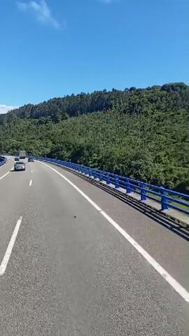 This media file captures a scenic drive on a highway, likely in Colunga, Spain. The video is shot from the perspective of a vehicle moving forward, offering a dynamic view of the road ahead and the surrounding landscape.

**Subjects:** The primary subjects are the highway itself, with its clear lanes and white markings, and the surrounding natural environment. Several vehicles are visible on the road, including cars and a white bus. There are no discernible people in a way that they are the focus, but they are present as occupants of the vehicles.

**Scene and Setting:** The setting is a multi-lane highway elevated on a bridge or viaduct. Blue guardrails line both sides of the roadway, separating it from the slopes below. The landscape is characterized by rolling hills covered in lush green trees, suggesting a temperate climate and perhaps a rural or semi-rural area. In the distance, mountains can be seen, partially shrouded in a light haze, indicating a vast and open expanse. The sky is a vibrant, clear blue with a few wispy white clouds, typical of a pleasant day.

**Activity or Event:** The media captures a continuous drive along the highway. The perspective indicates forward motion, with the road curving gently to the right. The other vehicles on the road are also moving, suggesting normal traffic flow.

**Notable Details:**
*   **Time of Day:** The bright sunlight and clear blue sky strongly suggest it is daytime, likely mid-morning to early afternoon.
*   **Weather:** The weather appears to be fair and sunny with good visibility.
*   **Location Cues:** The combination of the specific highway design (elevated with blue guardrails), the hilly, forested terrain, and the mention of Colunga, Spain, all contribute to a sense of place.
*   **Emotions:** The overall impression conveyed is one of a pleasant, relaxed journey through beautiful scenery. The clear weather and open road evoke feelings of freedom and perhaps a sense of adventure.

**Visible Text:** There is no legible text visible in this media file. The focus is entirely on the visual experience of the drive.