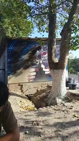 A man with dark curly hair, wearing a tan t-shirt, is sitting at a dark table outdoors. To his left, on the table, is an open, yellow beer can. In the background, there is a building with a large mural of two hands holding Pepsi cans. There are trees and a clear sky visible. A sidewalk with a few people and vehicles can be seen in the distance. The man is looking down and appears to be talking or reacting to something.