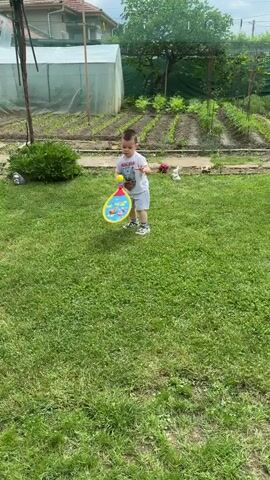 A young boy stands in a grassy backyard, holding a long yellow stick in his right hand. He wears a white graphic t-shirt, light gray shorts, and sneakers. Behind him to the left is a white greenhouse and a dark green bush. To the right, rows of garden plants are visible, along with small white decorative bird figures. The background features parts of a house roof and trees, under an overcast sky.
