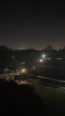 A nighttime video captures an elevated view of Rasoolpur, Pakistan, slowly panning from left to right. In the foreground, a large building with multiple windows and a distinct architectural style (reddish-brown with light-colored trim) is illuminated by several outdoor lights. Below it, a grassy area or courtyard is visible, with some benches or structures. The mid-ground features a landscape of trees and more buildings, interspersed with various city lights. In the far background, a prominent tall building stands out with bright lights near its top, suggesting a commercial establishment or hotel. The sky is dark, characteristic of nighttime, and there's a slight haze or dust in the air, causing the distant lights to appear somewhat diffused. No people or specific activities are visible; the video presents a static, panoramic view of the urban landscape at night.
