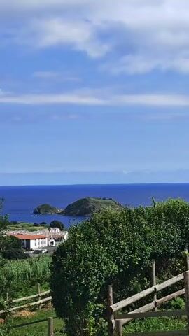 A scenic view overlooking a coastal village. In the foreground, there are lush green trees and a wooden fence. Beyond the trees, white buildings with red-tiled roofs are visible, nestled amongst vegetation. The village sits on the edge of a vibrant blue ocean. In the distance, a small, green island with a prominent peak rises from the water. The sky is a clear blue, dotted with fluffy white clouds, suggesting a sunny day. The overall impression is one of a peaceful, picturesque seaside landscape.