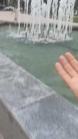 A close-up, shaky first-person perspective video shows a person's arm and hand near a fountain. The fountain has multiple spouts spraying water upwards, creating a curtain effect. The water is clear and turquoise in color. In the background, there are buildings and trees, suggesting an urban outdoor setting. The person's hand is extended towards the fountain, and the video appears to be capturing the interaction of the hand with the water spray.