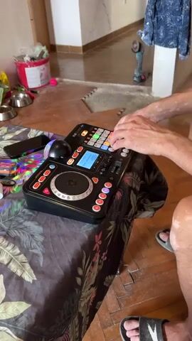 A person's hands are visible as they operate a black DJ controller with a circular illuminated disc and numerous buttons and a screen. The controller rests on a table covered with a floral patterned cloth. In the background, a partially constructed room is visible with exposed walls and floor tiling. There are also some household items such as buckets, pet bowls, and a phone on a shelf. The person is wearing casual clothing and sandals. The focus is on the DJ controller and the person's interaction with it.