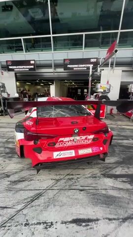 The video shows a race track pit lane with multiple race cars, including BMWs and a Porsche. The cars are various colors, including red, blue, and green. People, some in racing attire and some in casual clothing, are standing around the cars, talking and looking at their phones. The pit lane is wet and stained with tire marks. The sky is partly cloudy.