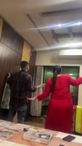 A man and a woman are standing in a room with their arms outstretched, mimicking airplane wings. The man is wearing a black and red plaid shirt and jeans. The woman is wearing a red dress. There is a yellow suitcase on the floor. The room has a wood floor and a ceiling fan. There is a large window in the background. A large wardrobe is against the left wall.