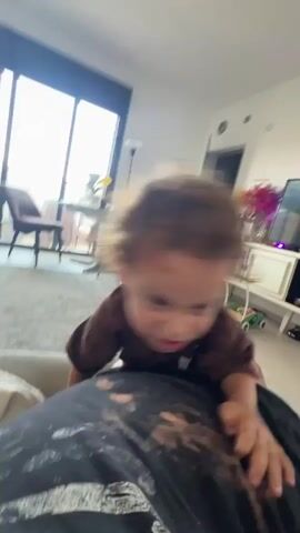 A child with curly brown hair wearing a brown t-shirt and white shorts is playing on a person lying on a couch. The child is looking at the camera and moving their hands. The background shows a window with natural light, a chair, a dresser with decorations, and a television. The camera moves erratically, at times blurring the image. The child appears to be interacting playfully with the person on the couch, with the video ending with the child embracing the person.