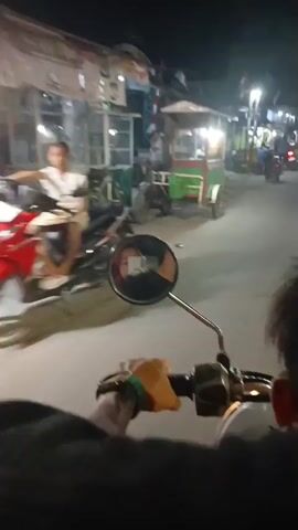 A first-person perspective shot from a motorcycle at night shows the handlebars and the rider's hand with a green wristband. In the mirror, a person wearing a mask and a headscarf is reflected. The motorcycle is moving down a street lined with buildings and roadside stalls, some of which are illuminated by lights. Other motorcycles are visible in the background, along with a few pedestrians. The street appears to be paved.