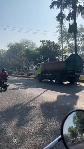 A dump truck is being driven down a paved road. There are several motorcycles on the road as well. The dump truck is green and has a gray bed. The motorcycles are various colors. The road is lined with trees and bushes. The sky is clear and blue.