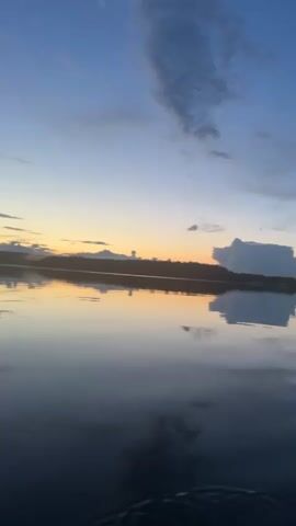 A video shows a serene body of water at dusk. The sky is a gradient of blues with touches of orange from the setting sun. Clouds are scattered across the sky, some catching the warm light. The water is calm, reflecting the sky and the distant tree line. A paddle is visible at the bottom right of the frame, suggesting the video was taken from a boat or kayak. The overall atmosphere is peaceful and tranquil.