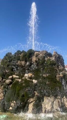 The video shows a close-up, low-angle shot of a large, rocky fountain with a powerful stream of water shooting high into the air. The rocks are covered in dark green moss and have lighter colored patches. Water cascades down the sides of the rock formation, creating a misty spray. The fountain is set against a clear, bright blue sky, indicating a sunny day. The overall impression is one of natural beauty and refreshment. There is no visible text in the video. The activity is the continuous operation of the fountain, with water being propelled upwards and falling back down. The scene is dominated by the fountain itself, with only a hint of buildings visible in the far background on the right side, suggesting an urban setting. The time of day appears to be daytime, given the bright sunlight and clear blue sky.