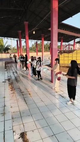 A group of young women, dressed casually, stand in a line under a covered outdoor area. They are holding circular objects, possibly fans or small drums, and appear to be engaged in a coordinated activity, possibly a dance or performance. The floor is tiled and appears to be somewhat damp or dirty in places. Several pink pillars support the roof of the structure. In the background, palm trees and buildings are visible, suggesting a tropical or subtropical location. The lighting indicates it is daytime, likely late afternoon due to the warm light.