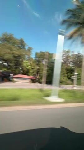 The video shows a view from inside a vehicle as it travels along a road. The scenery outside is blurred due to the motion, indicating the vehicle is moving at speed. The road appears to be paved, with a white line marking the edge. Trees and vegetation line the side of the road, and the sky is visible above, with some clouds. In one frame, a building with an orange roof is briefly visible, also blurred by the movement. The interior of the vehicle is partially visible, including a black seat and a metal bar with padding, suggesting it might be an SUV or a vehicle with a roll cage. The overall impression is that of a scenic drive through a wooded area.