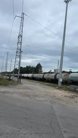 The video shows a stationary train consisting of tanker cars, several labeled "BULMARKET." The train is positioned on a track beside a paved road and gravel area. In the background are trees, and overhead are overcast skies and power lines supported by utility poles. There are no visible people or actions taking place.