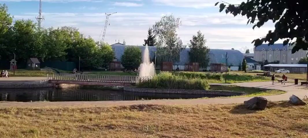 The video shows a park scene with a pond in the foreground and a fountain in the center. The fountain is spraying water high into the air, creating a mist that catches the sunlight. Around the pond, there are trees and grass. In the background, there are some buildings and a bridge. There are a few people visible in the park, some sitting on benches and others walking. One person is seen riding a bicycle. The sky is blue with some clouds. The overall scene is peaceful and idyllic.