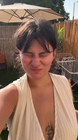 The video begins with a medium close-up shot of a woman's face. She has dark hair, some of which is falling forward onto her forehead, and she is wearing a light-colored, sleeveless top with pinstripes. A tattoo is visible on her chest. She is looking at the camera with a slight smile. The background appears to be an outdoor setting, possibly a backyard, with a thatched fence and an umbrella visible behind her. As the video progresses, water begins to fall from above, creating a "rain" effect. The woman's expression changes to one of surprise and amusement as the water hits her. She laughs and her hair becomes wet. The video continues with more shots of the woman in the "rain," with her expressions shifting between smiling, laughing, and looking directly at the camera. The water appears to be from a sprinkler or similar device. The video concludes with a close-up shot of the woman's face, her skin appearing flushed, and what look like blemishes or redness on her cheeks and jawline.