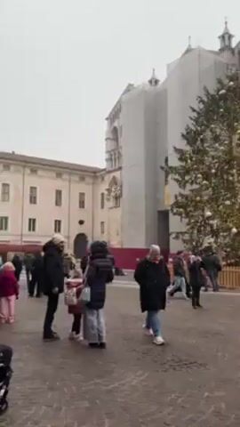 The video shows a town square in Italy, with a large Christmas tree decorated with gold balls as the central focus. There are several people walking around the square, most wearing dark winter clothing. Buildings surround the square, with an older stone building under scaffolding, and a more modern red brick building, which appears to house a McDonald's. The sky is overcast and grey, suggesting a cold day. A woman in a black coat and jeans walks towards the camera, then stops and looks to the left. A song is playing in the background.
