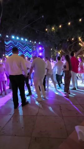 A group of people are dancing on a paved outdoor area at night.  A stage with a large screen displaying a zigzag pattern of blue and white lights is visible in the background. String lights are hung above the scene. The people are dressed in light-colored clothing, mostly white shirts and pants, with one person wearing a red shirt and another in a patterned dress. The lighting is primarily purple and blue, with some green light projected onto the dance floor. The overall atmosphere appears to be a celebration or party.