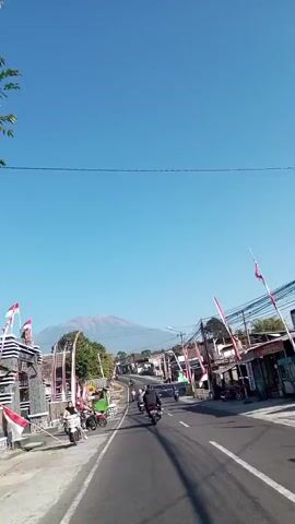 A vehicle-mounted camera perspective faces forward on a paved road, heading towards a distant, hazy mountain peak under a clear, bright blue sky. The road is lined with small buildings, many adorned with red and white striped flags. Several motorcycles are visible on the road, with some riders and passengers present. On the left side of the road, a few people are gathered near parked motorcycles. Utility poles with numerous wires run along the right side of the road. The overall scene suggests a warm, sunny day in a semi-rural or small town setting, possibly in Southeast Asia, given the presence of motorcycles and the style of the flags.