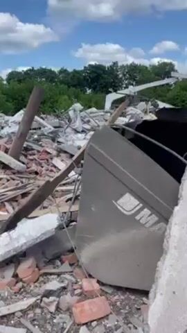 The video shows a close-up of the debris of a destroyed building. The wreckage consists of broken bricks, chunks of concrete, metal scraps, and twisted pipes. A gray metal panel with the word "MAKITA" on it is visible among the debris. In the background, a treeline and a partial structure of a building can be seen against a blue sky with clouds. The scene suggests destruction and the aftermath of an event that caused significant structural damage. The camera remains still throughout the video.